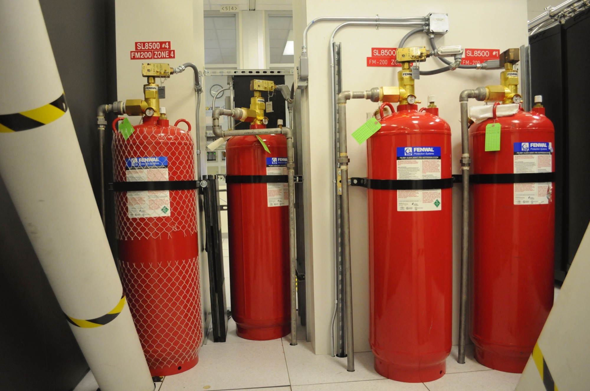 Four red clean-agent suppression cylinders standing against a wall in a server room, each with a brass discharge valve and pipework overhead.