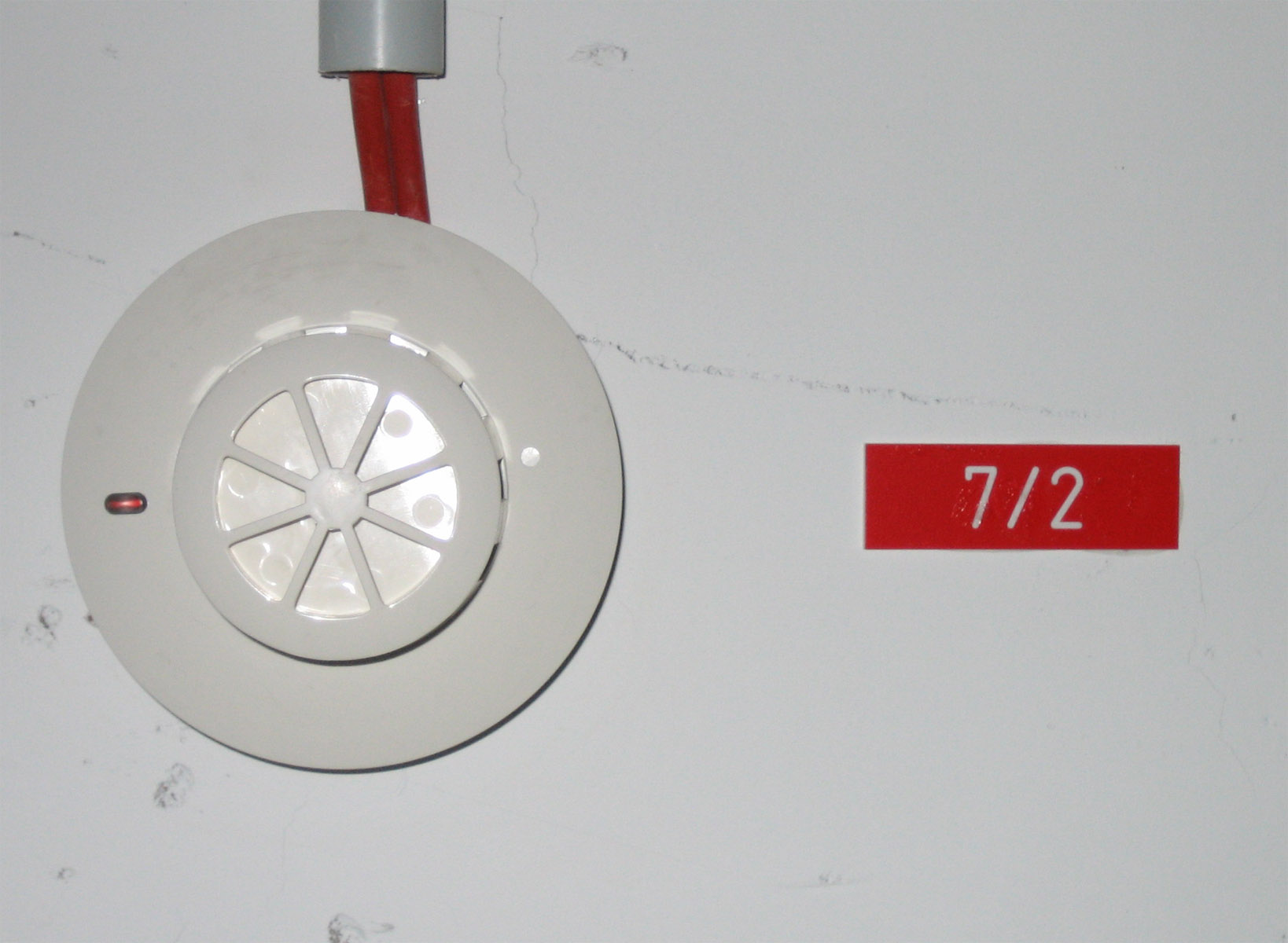 A white ceiling-mounted optical smoke detector with a red indicator lamp, beside a small red zone label reading 7/2.