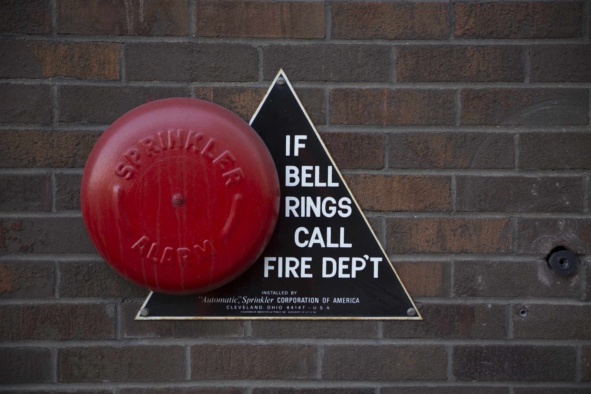 A red sprinkler alarm bell on a brick wall next to a sign reading "If bell rings call fire dept".