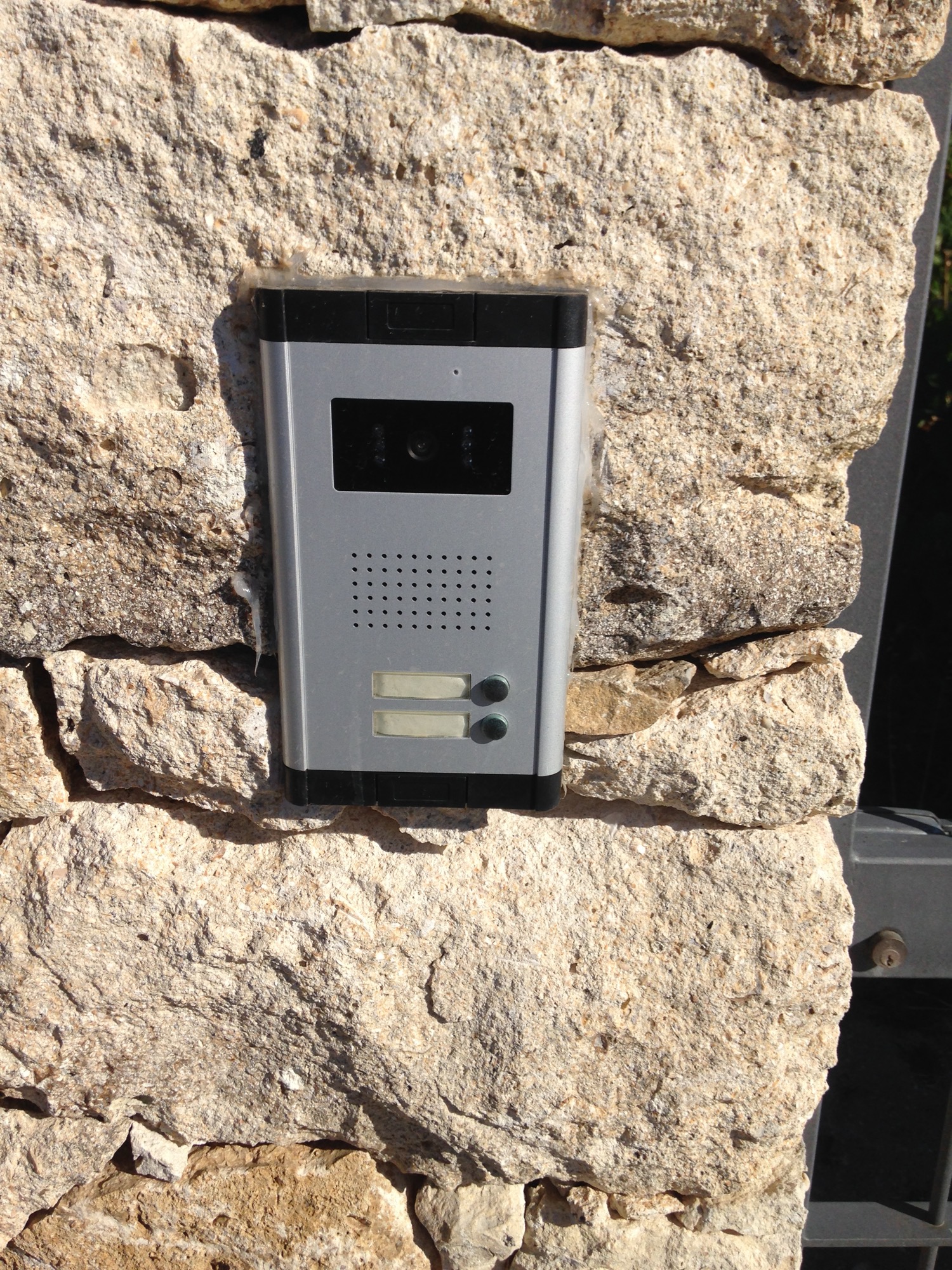 A two-button video door phone entry panel with camera and speaker, set into a stone wall beside a gate.